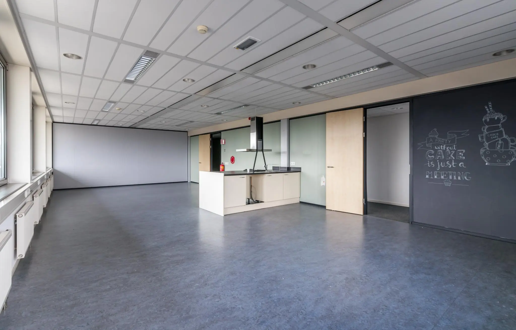 Leeg kantoorpand met een open keuken en een krijtbordmuur met de tekst “A party without cake is just a meeting”.